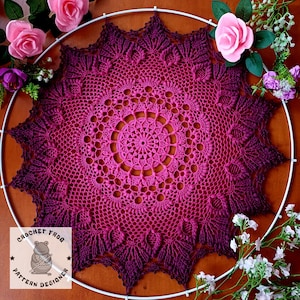 Könnte beinhalten: Eine große, gehäkelte Deckchen mit einem Farbverlauf von rosa und violetten Farbtönen. Das Deckchen ist rund und hat ein Sternmuster. Es wird auf einer Holzoberfläche mit Blumen drumherum gezeigt.