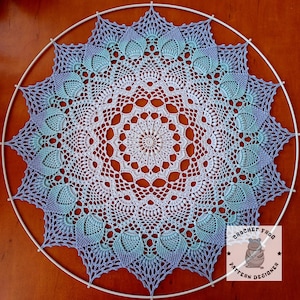 Op de afbeelding: Een handgemaakte, cirkelvormige gehaakte doily in tinten wit, lichtblauw en lavendel, gemonteerd op een witte metalen hoepel. De doily heeft ingewikkelde kanten patronen en een stervormig ontwerp. Het Crochet Frog Pattern Designer-logo staat rechtsonder.