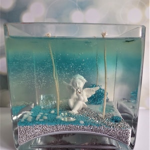 May include: A square glass candle holder filled with blue gel wax. Inside, a white angel figurine sits amidst blue and silver decorative elements. Two thin sticks extend upwards, and the candle holder has a silver base.