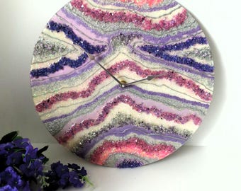 May include: A round clock with a white background and a colorful design of pink, purple, and silver glitter. The clock face is made of resin and has a unique, abstract design.