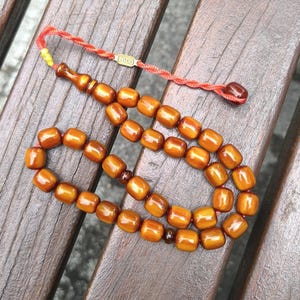 Op de afbeelding: Een bruine houten gebedsketting of rozenkrans met een rode kwast en een gouden label met de tekst "Agdo".