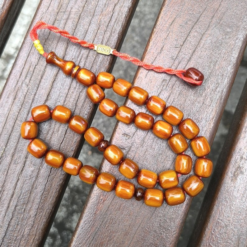Op de afbeelding: Een bruine houten gebedsketting of rozenkrans met een rode kwast en een gouden label met de tekst "Agdo".