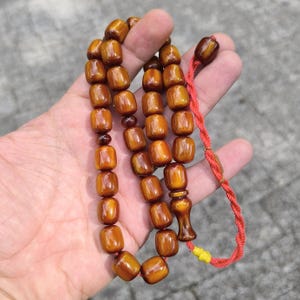 Op de afbeelding: Een bruine houten gebedsketting of rozenkrans met een rode kwast. De kralen zijn rond en hebben een gladde, gepolijste afwerking.