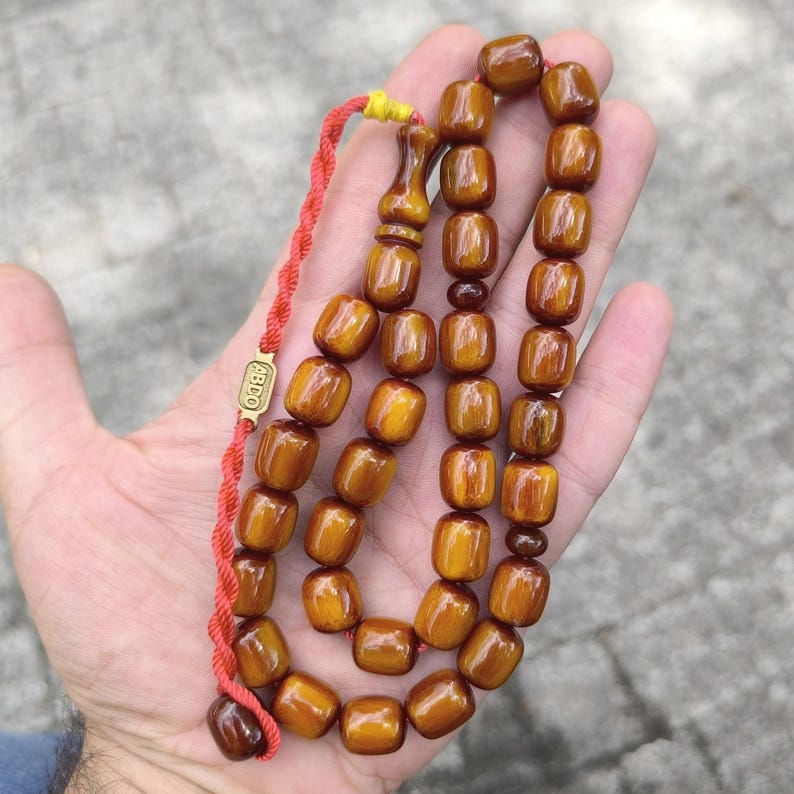 Op de afbeelding: Een bruine amber gebedskralen ketting met een gouden kwast en een rood touwtje. De kralen zijn rechthoekig en hebben een glanzende afwerking. De ketting is gelabeld met "ABDO".
