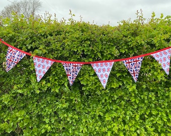 May include: A red, white, and blue bunting with a Union Jack pattern hangs against a green hedge. The bunting is made of fabric and has a red ribbon.