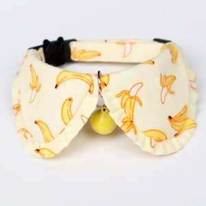 May include: A yellow and white cat collar with a bell. The collar has a ruffled edge and a pattern of yellow bananas on a white background.