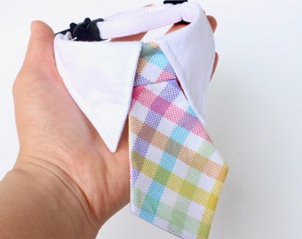 May include: A white fabric collar featuring a pointed collar design and an attached necktie. The necktie displays a multi-colored plaid pattern in shades of pastel pink, blue, yellow, and green. A black plastic buckle is visible on the collar strap.
