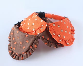May include: Two pet collars featuring ruffled fabric overlays. One collar is bright orange with a white pumpkin pattern, and the other is dark brown with an orange pumpkin pattern. Both collars include black plastic buckles and adjustable straps.