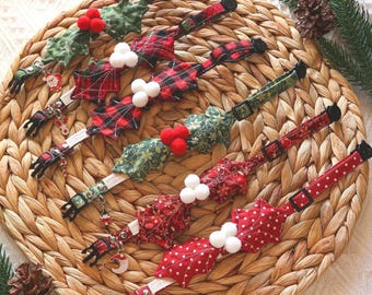 May include: A collection of five fabric pet collars featuring festive holiday designs. Each collar includes a decorative holly leaf attachment with small pom-poms and a dangling metal charm, such as a Santa, reindeer, or stocking.
