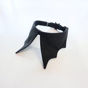 May include: A black bat-wing collar, likely for a pet or costume. The collar is made of a dark fabric and features a bat-wing design on each side. It has an adjustable strap with a metal buckle.