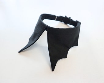 May include: A black bat-wing collar, likely for a pet or costume. The collar is made of a dark fabric and features a bat-wing design on each side. It has an adjustable strap with a metal buckle.