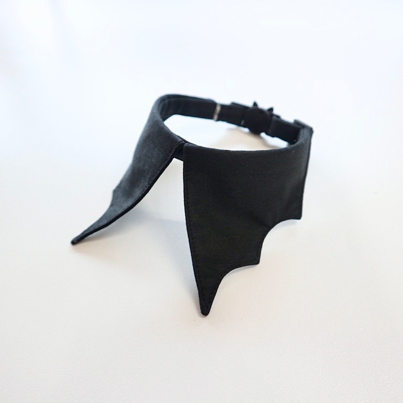 May include: A black bat-wing collar, likely for a pet or costume. The collar is made of a dark fabric and features a bat-wing design on each side. It has an adjustable strap with a metal buckle.
