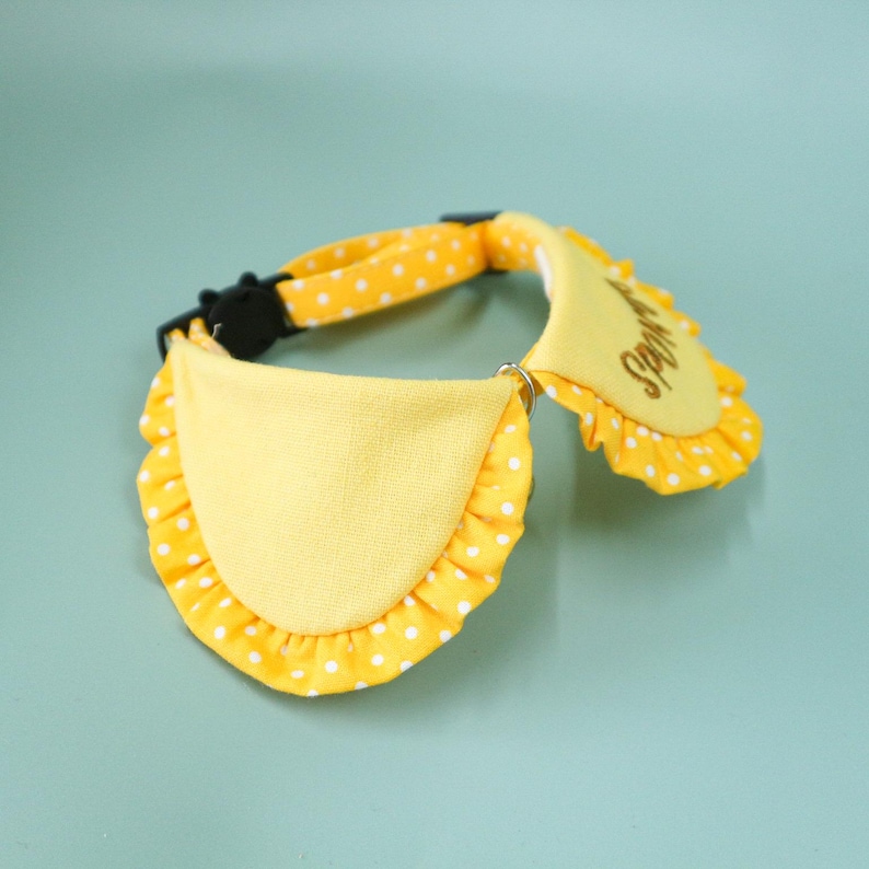 May include: A yellow pet collar with a matching bib. The collar is yellow with white polka dots and a black buckle. The bib is yellow with a ruffled edge and the word "Spoil" embroidered on it.