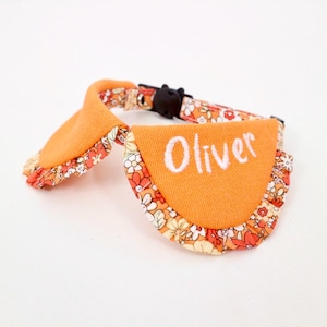 Puede incluir: Un collar para mascota con una sección de tela naranja vibrante bordada con el nombre Oliver en hilo blanco. El collar incluye un borde de tela con estampado floral en tonos naranja, rojo y crema, terminado con una hebilla de plástico negro.