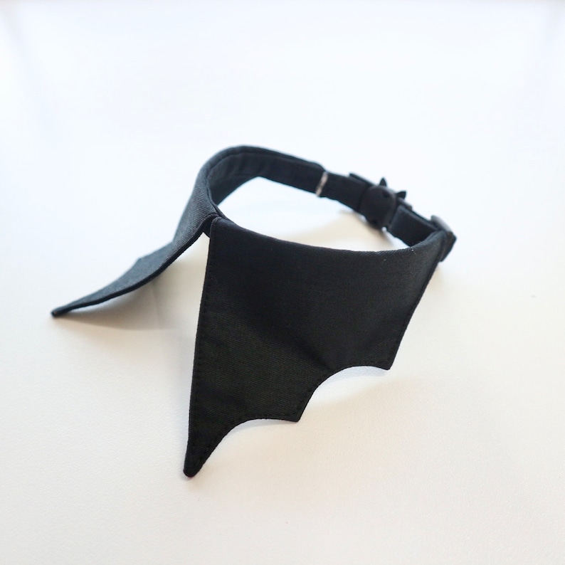 May include: A black bat-wing collar, likely for a pet. The collar is made of a solid black fabric and features a bat wing design on the front. It has a black adjustable strap with a silver-colored buckle.