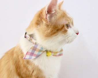 May include: A close-up of a ginger and white cat wearing a pink, yellow, and blue plaid bow tie with a bell.
