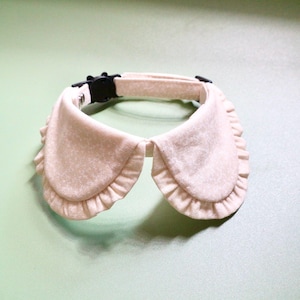 May include: A beige pet collar with a ruffled, decorative collar. The collar has a black plastic buckle and a soft, textured appearance. The design is reminiscent of a vintage or Victorian style.