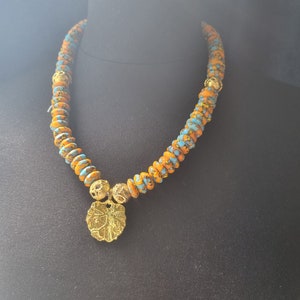 May include: A necklace featuring orange, blue, and yellow beads. It has a gold-coloured pendant with a floral design and gold-coloured accents. The necklace is displayed on a black mannequin.