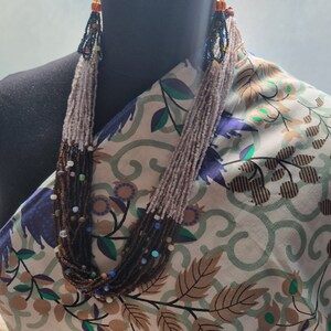 May include: A multi-strand beaded necklace in shades of brown, beige, and colourful beads. The necklace is displayed on a black mannequin and draped over patterned fabric with floral and leaf designs. The beads are small, creating a cascading effect.