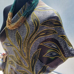 May include: A black mannequin draped with fabric featuring a leaf pattern in yellow, brown, and black. A multi-strand beaded necklace in shades of green and gold is around the neck. Beaded earrings are also visible.