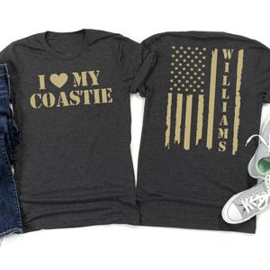 May include: Dark gray t-shirt with the text "I ♥ MY COASTIE" in gold lettering on the front. The back features a distressed American flag design with the name "WILLIAMS" in gold. A pair of gray sneakers is visible.