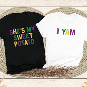 Op de afbeelding: Twee T-shirts met een bijpassend ontwerp. Het zwarte T-shirt zegt "She's my sweet potato" in groene en paarse letters. Het witte T-shirt zegt "I yam" in groene, gele en paarse letters.
