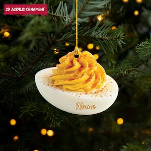 Peut inclure: Ornement de Noël en acrylique 2D en forme d'œuf farci, avec une garniture jaune et du paprika. Il est suspendu à un fil doré et le mot "Name" est écrit sur l'œuf. Le texte "2D Acrylic Ornament" est dans un rectangle rouge.