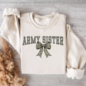 以下が含まれることがあります： クリーム色のスウェットシャツ。グレーのブロック体で「ARMY SISTER」の文字がプリントされ、その上に迷彩柄のリボンが描かれています。長袖でクルーネックです。