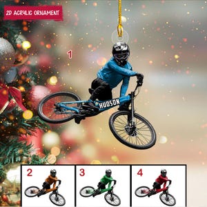 May include: A 2D acrylic ornament featuring a cyclist on a blue mountain bike with the name "HUDSON." The cyclist wears a blue jersey and helmet. The ornament is suspended by a gold string. Additional ornaments with cyclists in orange, green, and red are shown below.