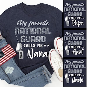 May include: Navy blue t-shirt with the text "My favorite NATIONAL GUARD calls me Nana" in white. The shirt is paired with blue jeans, white sneakers, and an American flag. Additional designs say "Papa", "Aunt", and "Uncle".