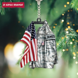 May include: A 2D acrylic ornament depicting the American flag and a military uniform with the name "JAMES." The ornament hangs from a silver chain and includes a dog tag with the American flag. The text "2D ACRYLIC ORNAMENT" is displayed at the top.