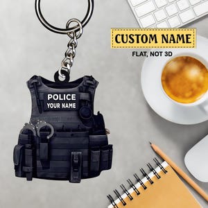 Pode incluir: Um chaveiro de colete policial preto com acessórios prateados. O chaveiro apresenta o texto "POLICE" e "YOUR NAME" em um fundo branco. O texto "CUSTOM NAME" também é visível. O chaveiro inclui algemas e outros acessórios.