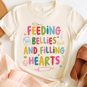 以下が含まれることがあります： カラフルな文字で「FEEDING BELLIES AND FILLING HEARTS」と書かれた白いTシャツ。テキストは、牛乳パック、サンドイッチ、カップケーキ、麺棒、リンゴなど、さまざまなカラフルな食べ物のイラストで囲まれています。