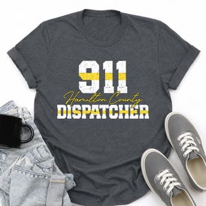 May include: A dark charcoal gray t-shirt featuring the white text 911 with a horizontal yellow line through the numbers, followed by the cursive yellow text Hamilton County and the block white text DISPATCHER with a yellow line through it.