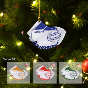 May include: A white and blue hockey goalie glove ornament with the name "Joseph" printed on it. The ornament is hanging on a Christmas tree.