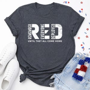 Könnte beinhalten: Ein dunkelgrau meliertes Kurzarm-T-Shirt mit dem weißen, abgenutzten Schriftzug REMEMBER EVERYONE DEPLOYED und dem kleineren Text UNTIL THEY ALL COME HOME auf der Brust. Das T-Shirt hat einen Rundhalsausschnitt und umgeschlagene Ärmelbündchen.