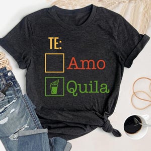 May include: A dark grey t-shirt with the text "TE: Amo Quila" in red, yellow, and green. The text is in a box with a tick next to it.