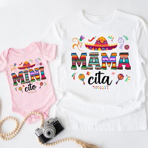 以下が含まれることがあります： 白のTシャツとピンクのベビーボディスーツ。どちらもカラフルな文字とソンブレロのデザインが施されています。Tシャツには「Mama cita」、ボディスーツには「Mini cito」と書かれています。マラカスと花のアクセントも付いています。