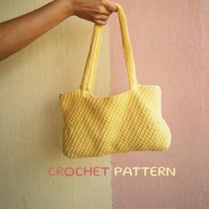 May include: A yellow crocheted tote bag with a textured pattern. The bag has two straps and is hanging from a hand.