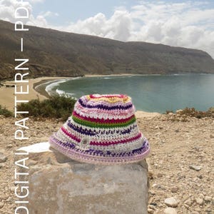 Peut inclure: Un chapeau seau crocheté multicolore avec des rayures horizontales roses, violettes, vertes et jaunes. Le chapeau est posé sur une pierre claire, avec un paysage côtier en arrière-plan. Le texte "DIGITAL PATTERN - PDF" est visible.