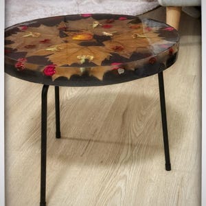 Puede incluir: Una mesa auxiliar redonda con estructura y patas de metal negro. La tapa es de resina transparente, que encierra hojas secas y pequeños pétalos de flores rojas y amarillas. La mesa tiene aproximadamente 46 cm de diámetro.