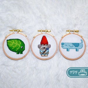 Puede incluir: Tres aros de bordado con diseños de punto de cruz. El primer aro presenta una lima verde, el segundo aro presenta un gnomo con un sombrero rojo, y el tercer aro presenta un perro azul.