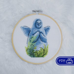 Peut inclure: Broderie au point de croix d'une figure sereine dans des tons bleus et verts, encadrée dans un cercle en bois. Le motif représente une figure aux mains jointes, entourée de verdure. Icône PDF en bas à droite.