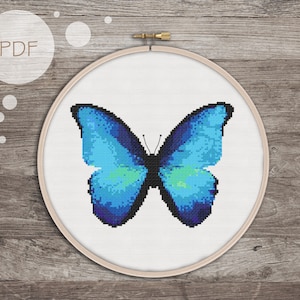 May include: A blue butterfly cross stitch pattern in a white embroidery hoop. The butterfly has a blue and green gradient with black outlines.