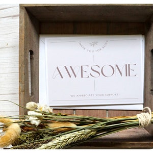 May include: A wooden crate holds a stack of white cards with the word "AWESOME" in mauve lettering. The cards also read "THANK YOU FOR BEING" and "WE APPRECIATE YOUR SUPPORT!". A bundle of dried flowers and grasses rests in front of the cards.