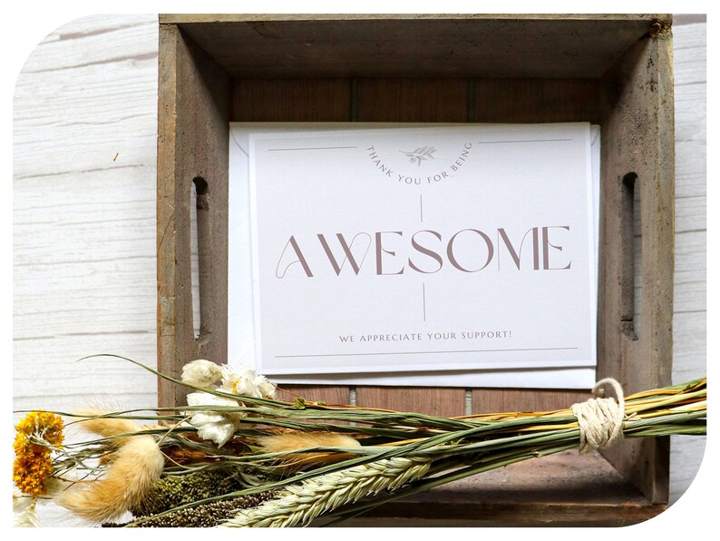 May include: A wooden crate holds a stack of white cards with the word "AWESOME" in mauve lettering. The cards also read "THANK YOU FOR BEING" and "WE APPRECIATE YOUR SUPPORT!". A bundle of dried flowers and grasses rests in front of the cards.