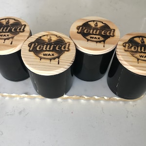 May include: Four black candles with wooden lids. Each lid has a wood-burned design of a record with the text "Poured Wax" on it.