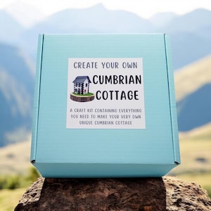 May include: A light blue box with a white label that says "Create Your Own Cumbrian Cottage." The label also includes a small illustration of a cottage and the text "A craft kit containing everything you need to make your very own unique Cumbrian Cottage."