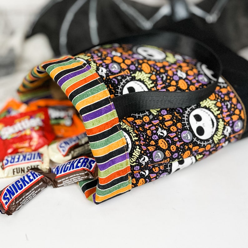 Puede incluir: Una bolsa de tela para truco o trato de color negro y verde con un forro a rayas y un asa negra. La bolsa est&aacute; llena de barras de caramelo, incluyendo Snickers y Fun Size.