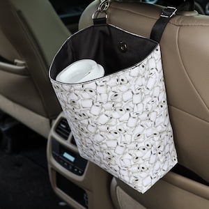 Puede incluir: Una bolsa de basura para coche blanca con una parte superior negra y una correa negra. La bolsa está colgada en el respaldo de un asiento de coche. La bolsa está cubierta con un patrón de rollos de papel higiénico.